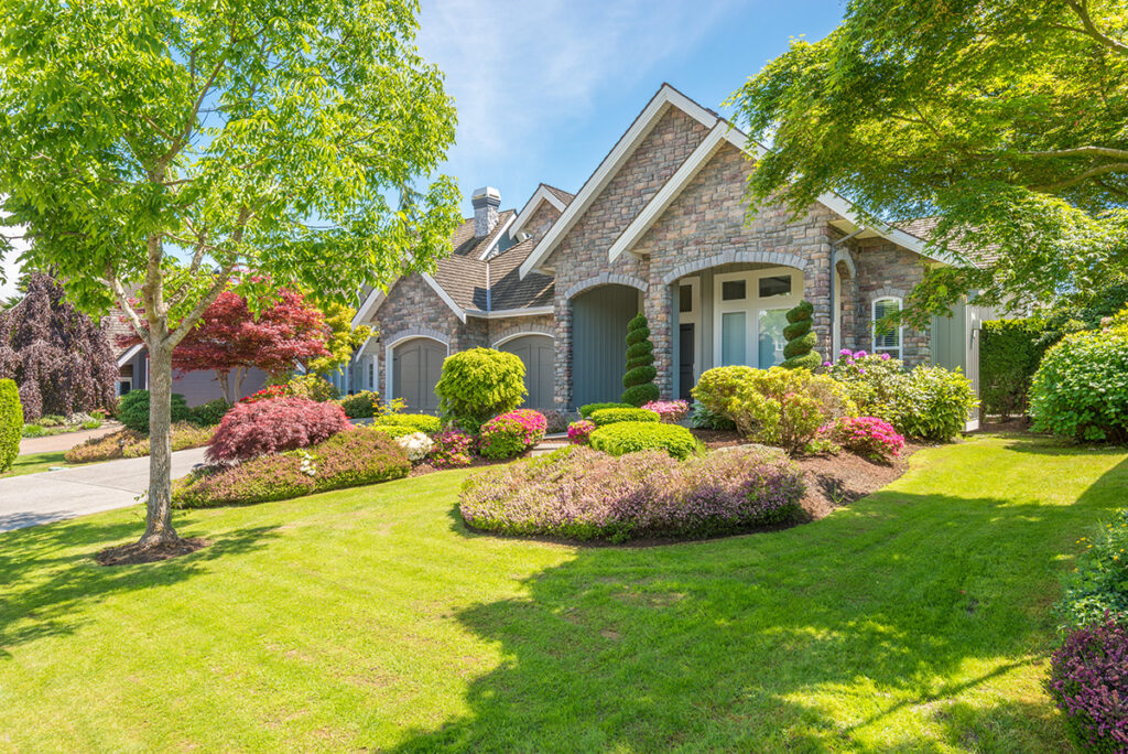 A brick house on a large high-value property with a green front lawn and lots of trees against a blue sky