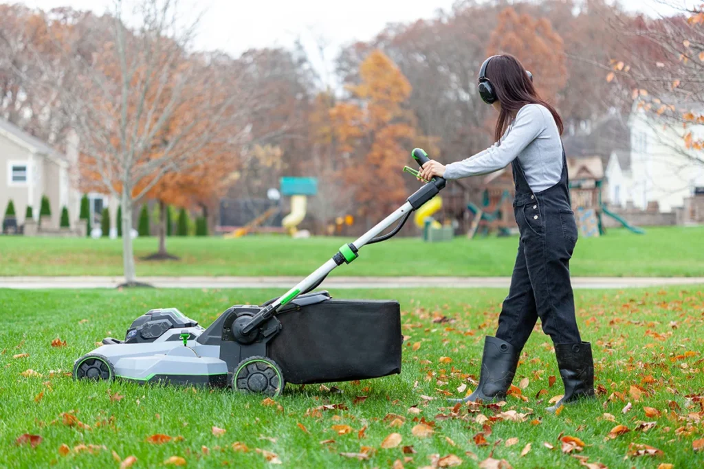 A woman wearing headphones uses a mulch mower to manage fallen tree leaves in her front lawn